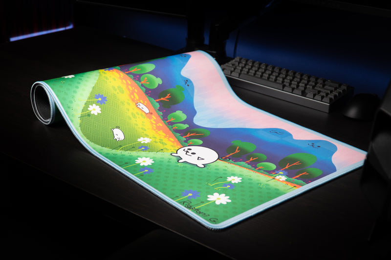Pippa Peep's Pipaland deskmat partially rolled on a dark desk, displaying flowers, hills, and the stitched edge.