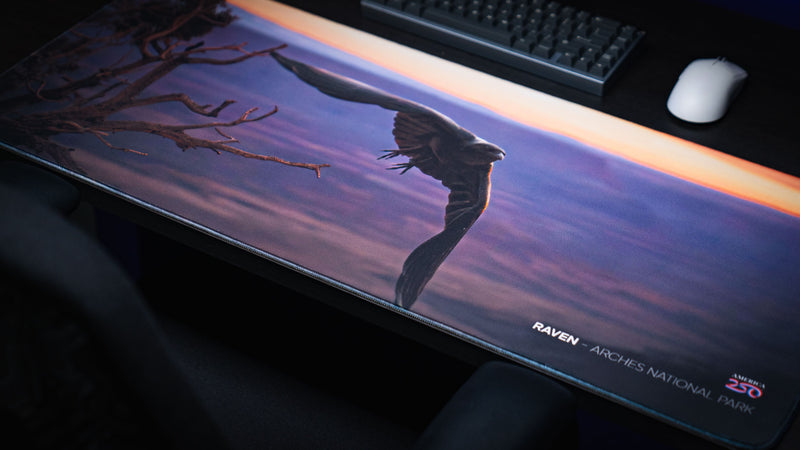 Angled view of the Raven America 250 deskmat featuring a raven in flight above Arches National Park at sunset with a premium gaming desk setup.
