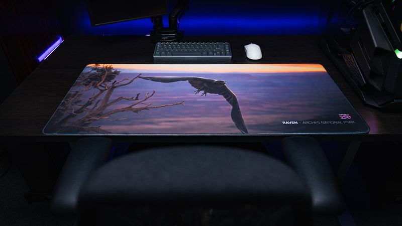 Front view of the Raven America 250 deskmat in a dark gaming setup with a raven in flight, Arches National Park sunset tones, keyboard, and white mouse.