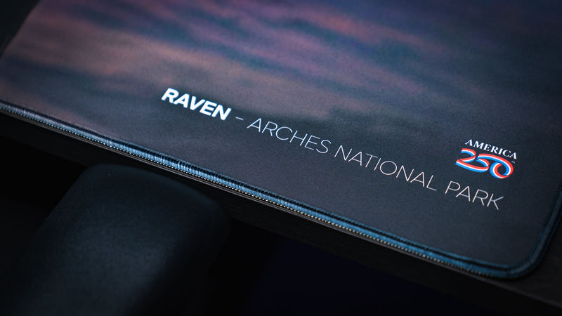 Close-up of the Raven America 250 deskmat showing Arches National Park title text, America 250 logo, dark canyon artwork, and stitched edge.