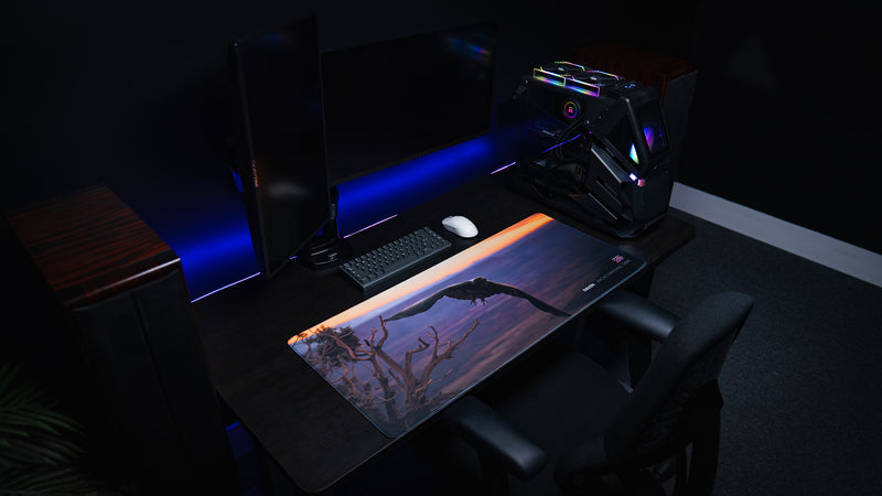 Premium Raven deskmat displayed on a black gaming desk with dual monitors, RGB PC, mechanical keyboard, white mouse, stitched edges, and dramatic purple sunset artwork from Arches National Park.