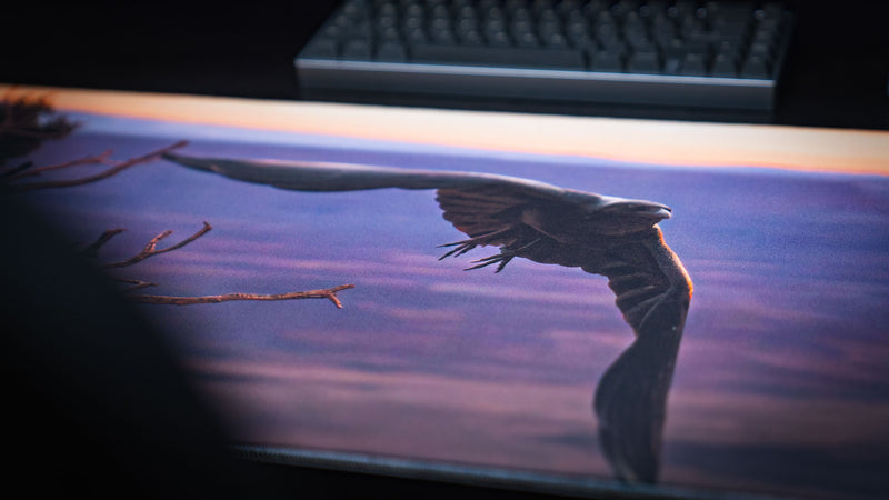 Macro view of the Arches National Park Raven mousepad showing a flying raven, desert tree branches, warm sunset glow, and premium stitched gaming deskmat surface.