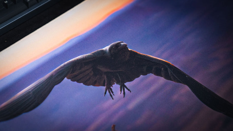 Macro view of the Raven America 250 deskmat showing a raven in flight across purple canyon tones with warm sunset light and detailed fabric texture.