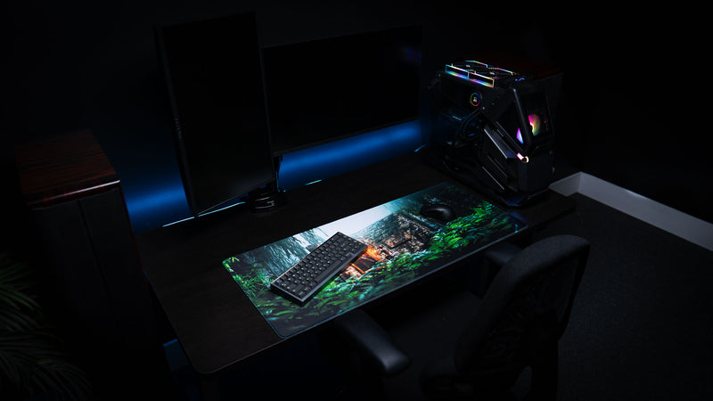 The Doorcamper / Angle 1 Rust deskmat displayed in a modern gaming setup, featuring a jungle stone base ambush scene with vibrant foliage, a mechanical keyboard, gaming mouse, and RGB PC.