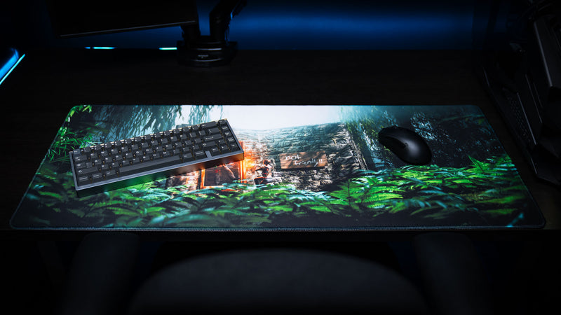 The Doorcamper / Angle 1 Rust deskmat displayed with a compact mechanical keyboard and gaming mouse, featuring a tropical stone base ambush scene with dense jungle foliage and premium stitched edges.