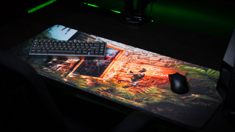 The Doorcamper / Angle 2 Rust deskmat displayed in a premium gaming setup with a mechanical keyboard and mouse, featuring a dramatic stone base ambush, dense jungle foliage, and high-detail printed artwork