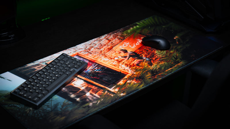 The Doorcamper / Angle 2 Rust deskmat displayed with a mechanical keyboard and gaming mouse, featuring an intense stone base firefight, lush tropical foliage, and premium stitched-edge construction.
