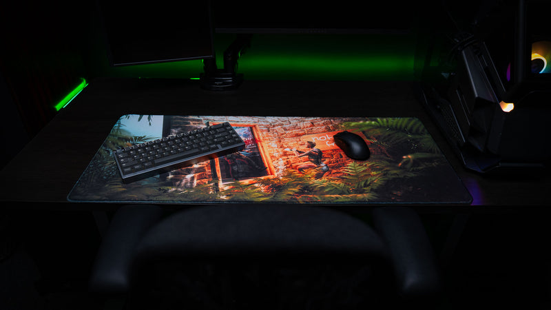 The Doorcamper / Angle 2 Rust deskmat displayed in a gaming setup with a compact mechanical keyboard and gaming mouse, showcasing a tense jungle base ambush scene with premium stitched edges and vibrant artwork.