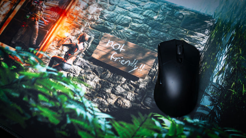 Close-up of The Doorcamper / Angle 1 Rust deskmat featuring a stone base entrance with a "Not Friendly!!" warning sign, armed survivors, and lush jungle foliage alongside a gaming mouse.