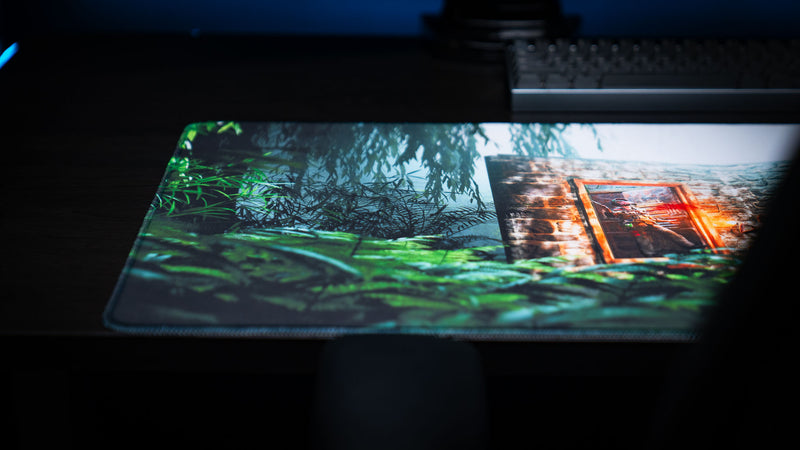 The Doorcamper / Angle 1 Rust deskmat featuring a close-up of a jungle stone base entrance framed by dense tropical foliage, showcasing vibrant colors and premium stitched edges on a gaming desk.
