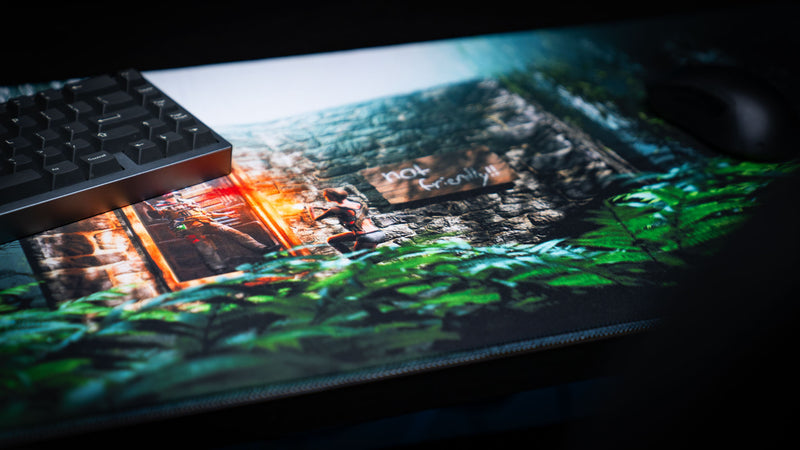 The Doorcamper / Angle 1 Rust deskmat featuring a jungle-surrounded stone base, with a survivor guarding the entrance and detailed tropical foliage, shown in a premium gaming desk setup.