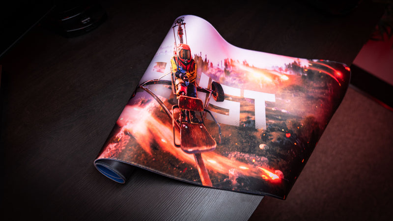Rolled corner close-up of the Rust “Minicopter Joyride” gaming deskmat showcasing cinematic helicopter artwork and stitched edges on a dark desk.