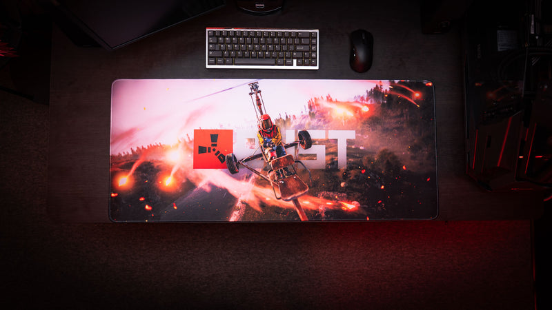 Full gaming desk setup featuring the Rust “Minicopter Joyride” deskmat with cinematic aerial combat artwork, keyboard, and mouse on a dark desktop.