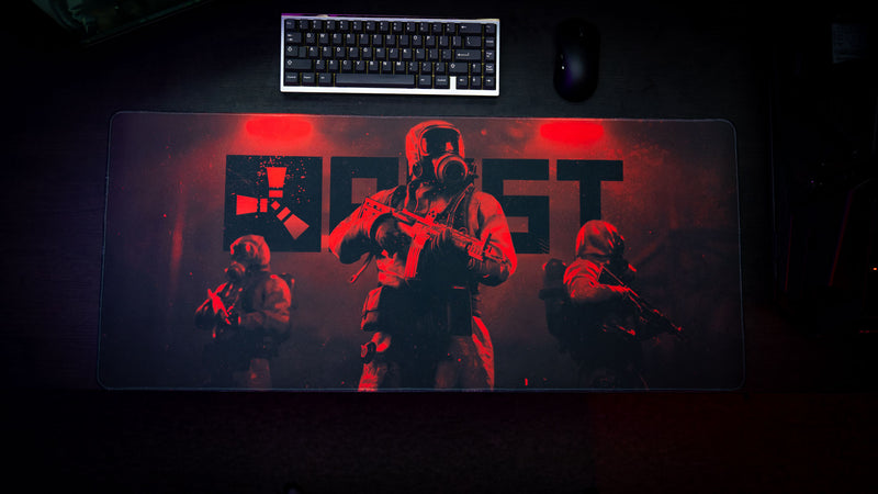 Top-down view of the Rust Scientist Trio deskmat featuring three armed scientists, the iconic Rust logo, dramatic red lighting, and a premium gaming setup.