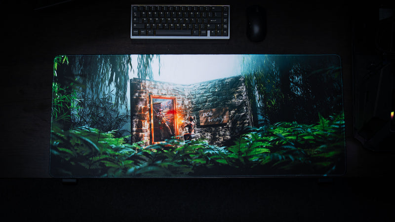 The Doorcamper / Angle 1 Rust deskmat displayed on a gaming desk, featuring a jungle-surrounded stone base with a heavily armed survivor waiting outside the doorway in a tense ambush scene.