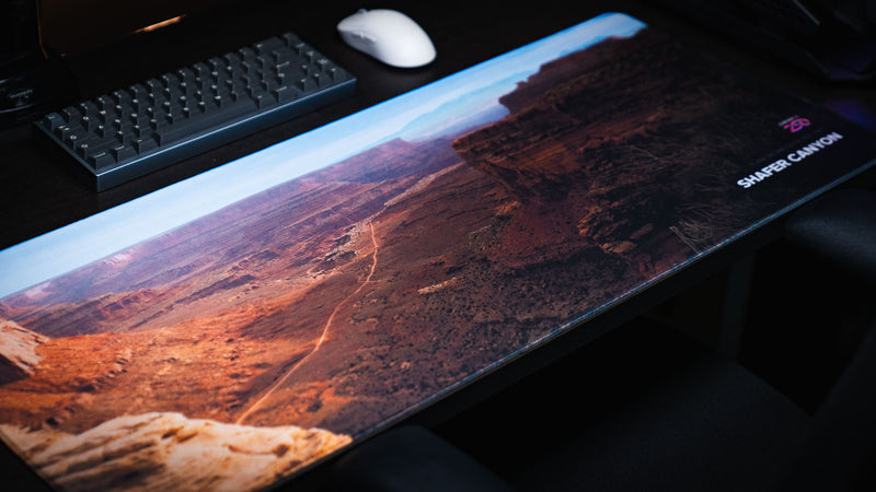  Shafer Canyon deskmat displayed on a dark desk with red rock canyon scenery, distant desert mountains, America 250 branding, and stitched edge.