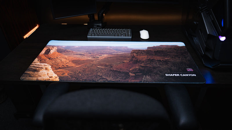 Front workstation view of the Shafer Canyon America 250 deskmat featuring Canyonlands National Park red rock cliffs, desert valley roads, keyboard, and mouse.