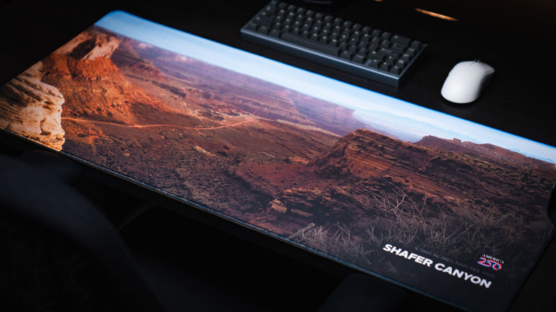 Shafer Canyon America 250 deskmat shown with keyboard and mouse, featuring Canyonlands desert cliffs, layered mesas, and warm red rock scenery.