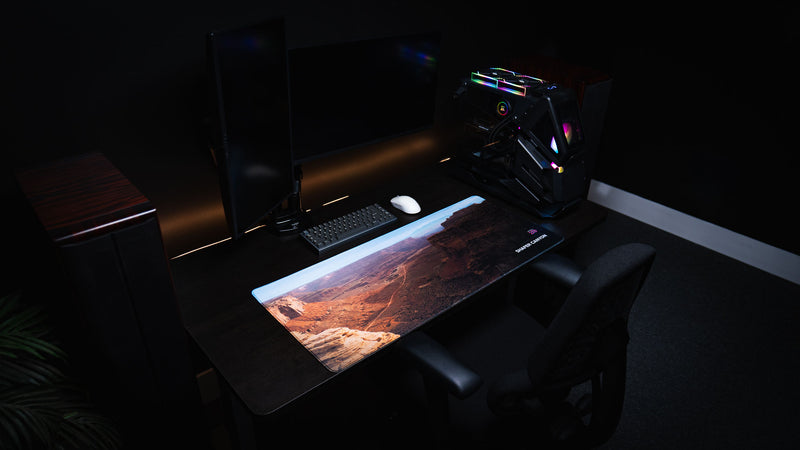 Left-angle view of the Shafer Canyon America 250 deskmat featuring Canyonlands red rock scenery, keyboard, mouse, PC, and dark desk setup.