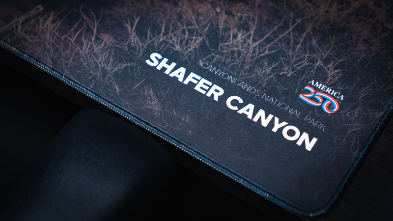 Shafer Canyon deskmat detail featuring Canyonlands National Park branding, dark desert texture, America 250 logo, and premium stitched border.