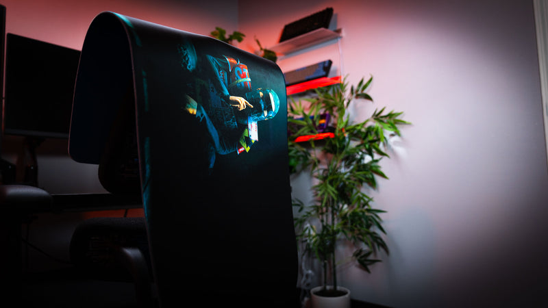 Shh Rust deskmat draped over a gaming chair, showcasing the dark minimalist artwork of a masked figure making a silence gesture with premium stitched edges and vibrant print quality.