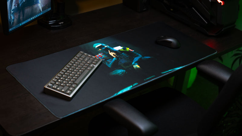 The Shh Rust deskmat displayed across a gaming desk with keyboard and mouse, featuring the iconic shushing character artwork illuminated against a dark background.