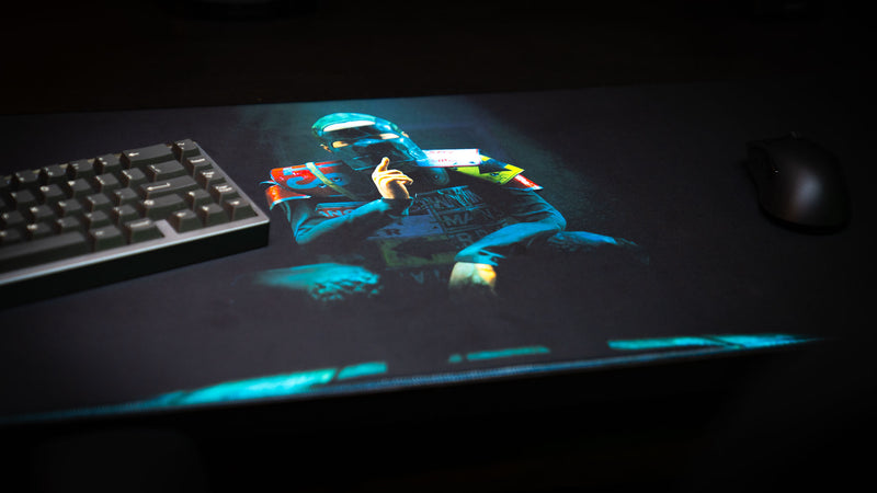 The Shh Rust deskmat displayed in a gaming setup with keyboard and mouse, showcasing the mysterious masked character artwork against a dark, atmospheric background.
