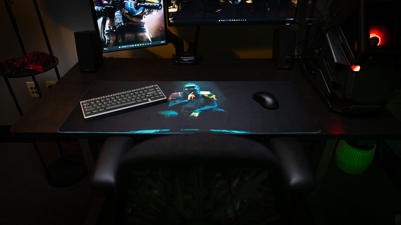 Shh Rust deskmat showcased in a gaming setup with keyboard and mouse, featuring a spotlight-lit masked figure making a silence gesture against a dark atmospheric background.
