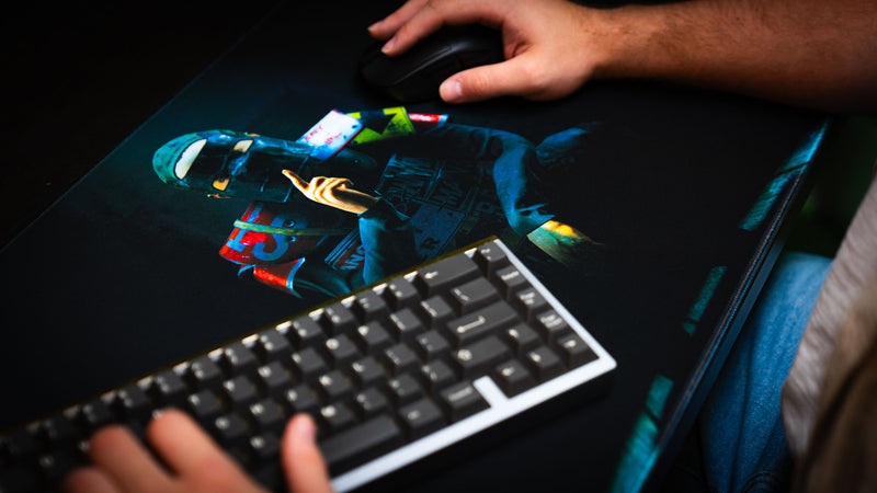 Player using the Shh Rust deskmat with keyboard and mouse, highlighting the detailed artwork of a masked figure making a silence gesture in a dark cinematic setting.