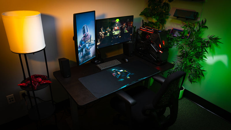 The Shh Rust deskmat displayed in a complete gaming setup with dual monitors, custom PC, ambient lighting, and the masked character artwork centered across the premium XXL mousepad.