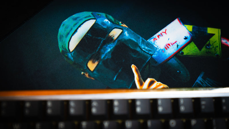 Detailed close-up of the Shh Rust deskmat featuring the iconic masked character making a shushing gesture, viewed from behind a keyboard in a gaming setup