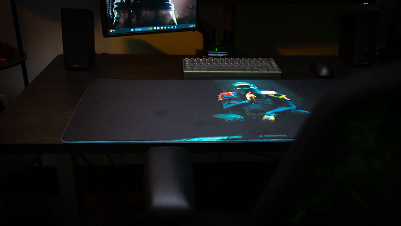 Shh Rust deskmat displayed on a gaming desk, featuring a spotlight-lit masked figure making a silence gesture against a dark minimalist background with stitched edges.