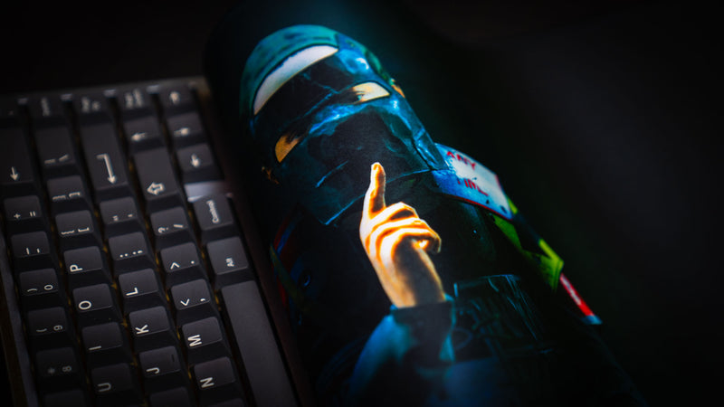 Close-up of The Shh Rust deskmat rolled beside a mechanical keyboard, showcasing the masked character artwork, vibrant colors, and premium stitched-edge construction.