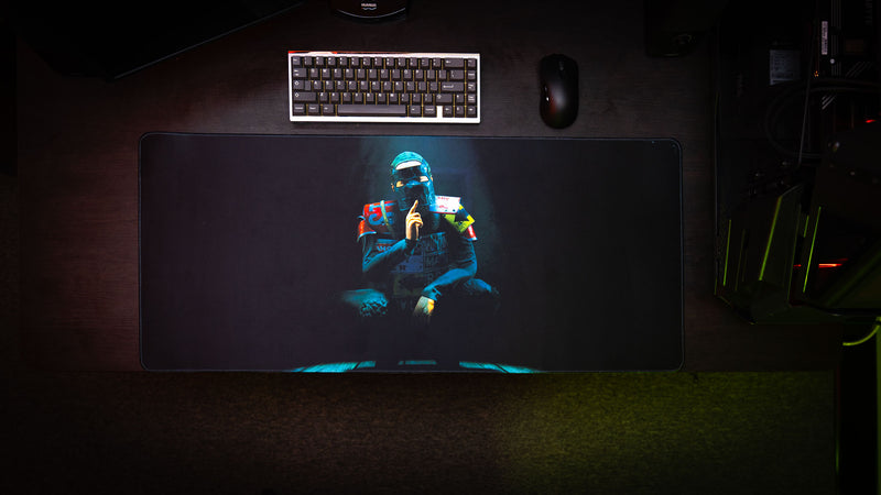 Top-down view of the Shh Rust deskmat featuring a masked figure making a silence gesture beneath a spotlight, displayed in a gaming setup with keyboard and mouse.