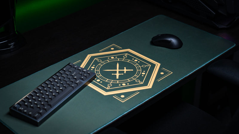 Angled close-up of the gold crossed-swords emblem on House Veridian deskmat beside a keyboard and mouse