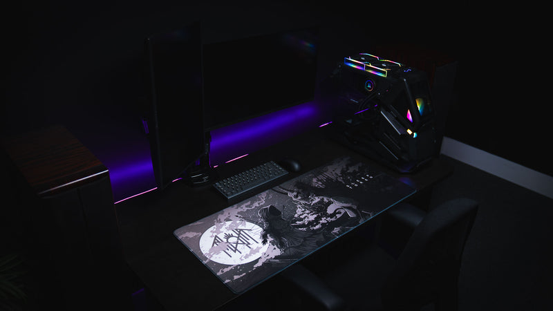 Wide gaming setup with The Battle deskmat below dual monitors and a lit PC