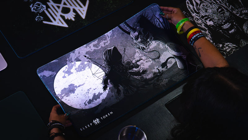 Angled overhead view of a player holding The Battle Sleep Token playmat, showing the moon, hooded figure, and creature