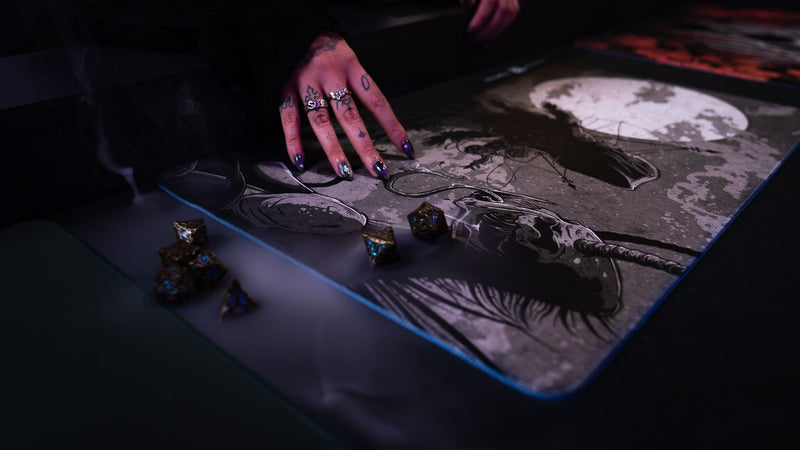 Close-up of The Battle Sleep Token playmat with a player's hand and gaming dice beside its moonlit artwork