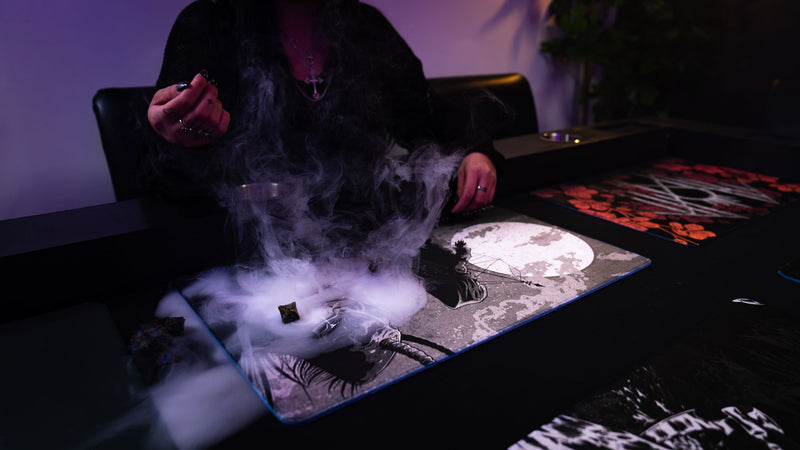 The Battle Sleep Token playmat on a gaming table with a player and drifting mist over its moonlit design