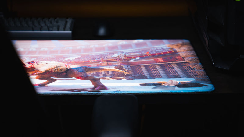 Side-angle view of The Spinosaurus Chompers deskmat by Keenling showcasing the Roman gladiator arena backdrop and the fierce dinosaur battle artwork with premium stitched edges.