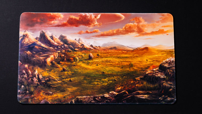 Fantasy landscape playmat featuring towering mountain peaks, rolling golden plains, distant hills, scattered trees, and glowing sunset clouds over a vast wilderness valley.