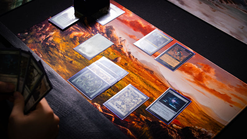 Sunspire Plains fantasy playmat in use during a tabletop card game, featuring golden grasslands, rocky peaks, and dramatic sunset skies.