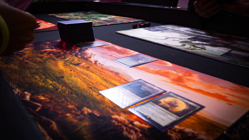 Sunspire Plains fantasy playmat in use during a tabletop card game, featuring rolling grasslands, rocky terrain, and a golden sunset horizon.