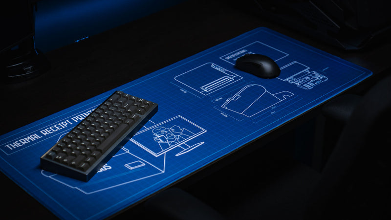 Angled view of the Thermal Receipt Printer blueprint deskmat inspired by Rust, featuring detailed technical schematics beneath a compact mechanical keyboard and gaming mouse on a premium extended gaming mouse pad.