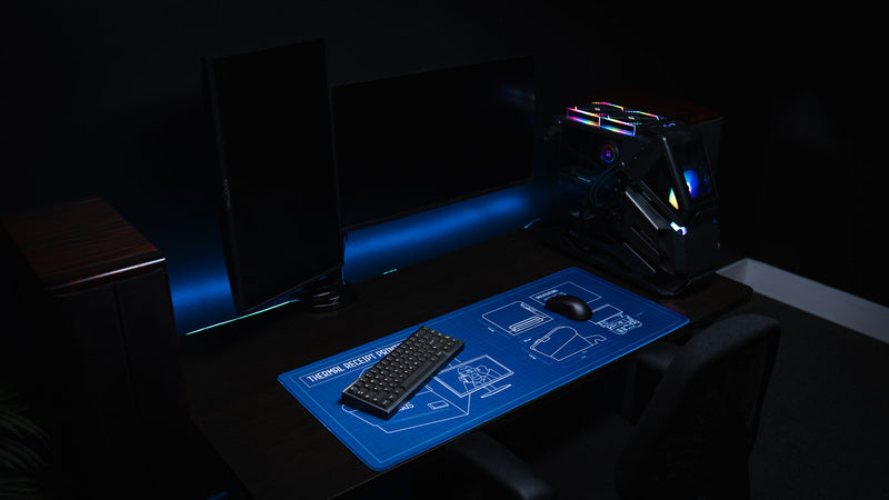 Minimal gaming workstation featuring the Thermal Receipt Printer blueprint deskmat inspired by Rust, displayed with a mechanical keyboard, gaming mouse, and RGB PC, showcasing the technical blueprint design across the full desk mat.