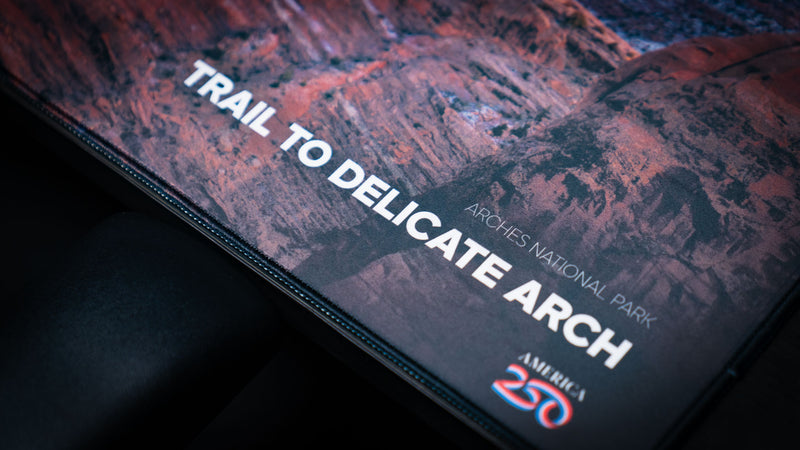 Close-up of the Trail to Delicate Arch deskmat featuring the Arches National Park title, America 250 logo, premium stitched edges, and detailed red rock photography.