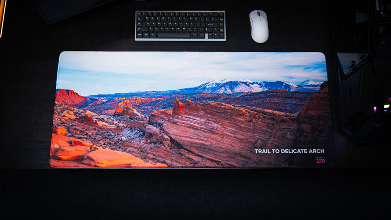 Trail to Delicate Arch deskmat displayed in a desktop gaming setup, showcasing the iconic sandstone trail, expansive red rock landscape, and distant snow-capped mountains of Arches National Park.