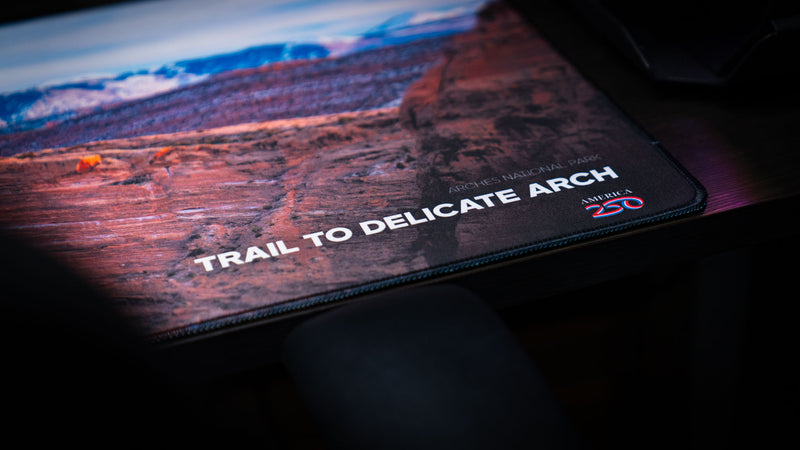 Close-up of the Trail to Delicate Arch deskmat highlighting the Arches National Park branding, America 250 logo, premium stitched edges, and crisp.