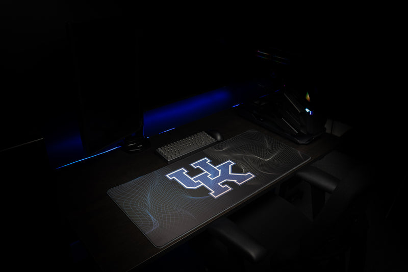 Kentucky Afterglow deskmat shown at an angle in a dark gaming setup with a glowing blue UK logo.