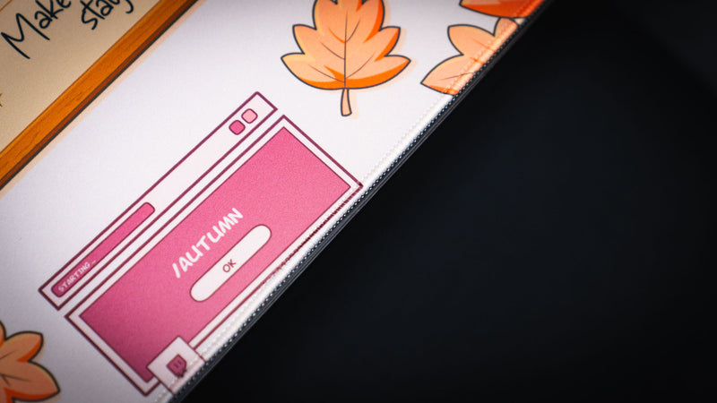 Close-up of the Welcome to the Leaf Pile deskmat featuring a pink autumn-themed desktop window illustration with falling maple leaves and stitched edge detail.